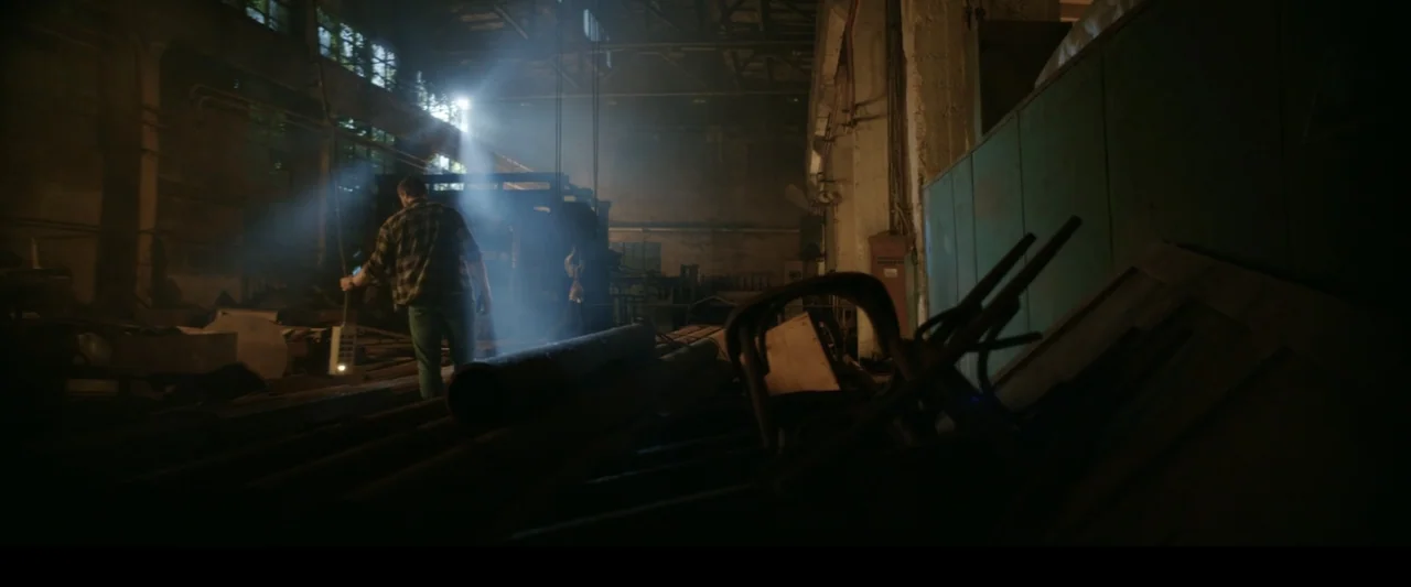 Black Lotus (2023) movie still: wide — An industrial facility interior filled with scattered metal pipes and debris under dusty light. A man stands…