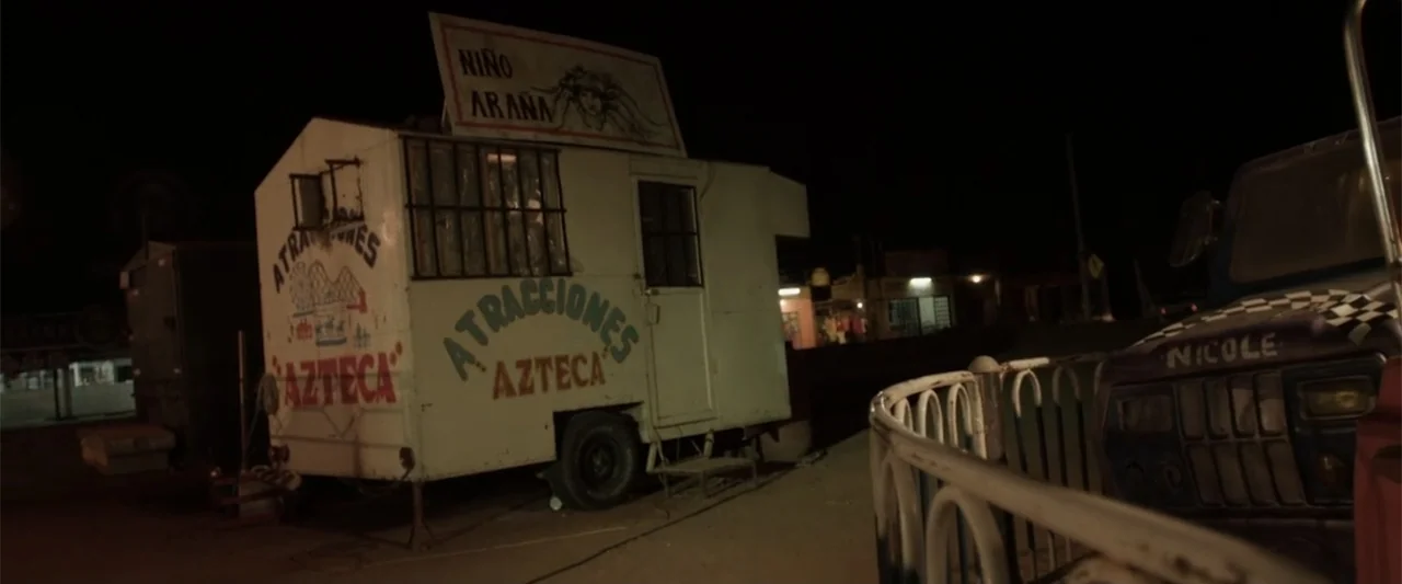 At Dawn (2013) movie still: wide — A collection of weathered carnival trailers sits in a dark, empty dirt lot at night. The white metal trailers…