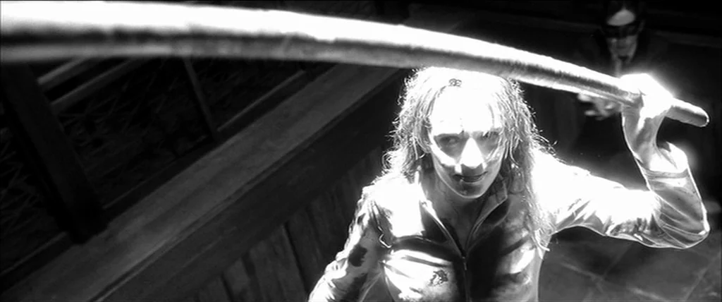 Kill Bill Vol. 1 (2004) movie still: medium — A dark, narrow corridor with wood paneling and a blurred secondary figure behind. A woman looks up…