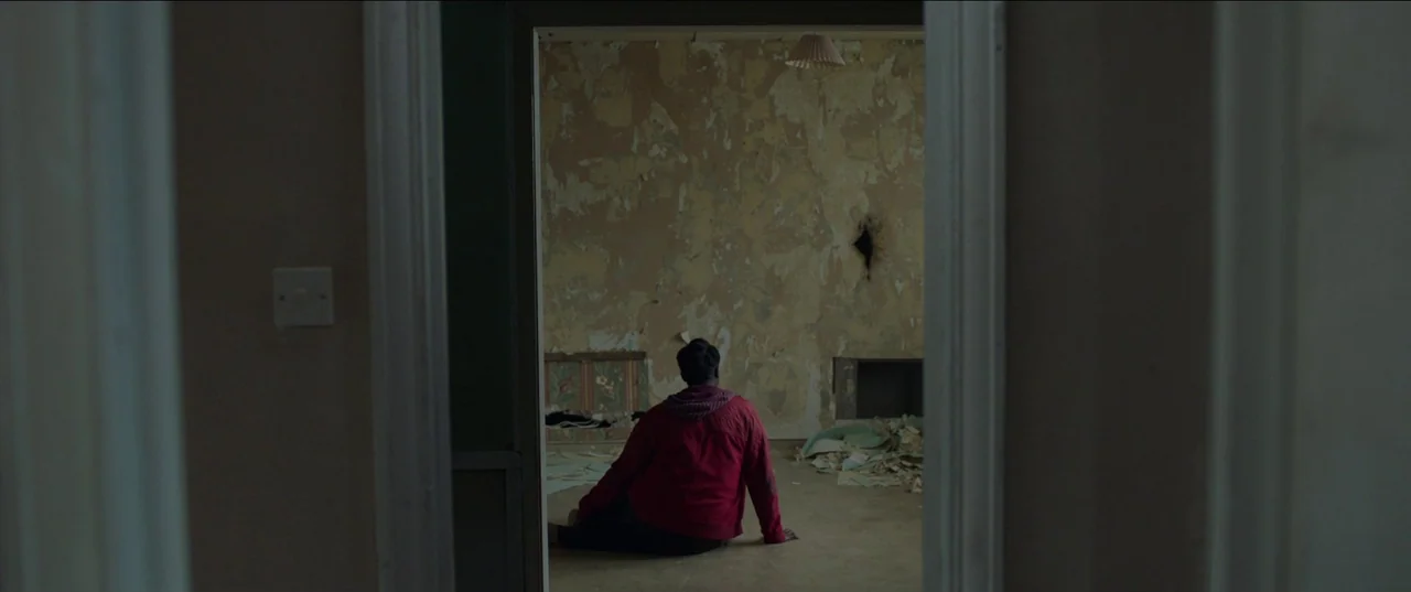 His House (2020) movie still: wide — A dilapidated interior space features peeling wallpaper and scattered debris on the floor. A person in a red…