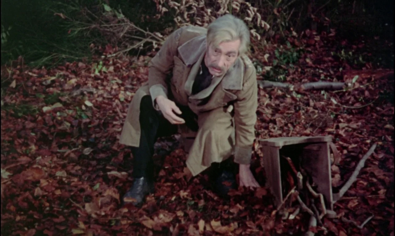 The Curse of Frankenstein (1957) movie still: medium — An elderly man kneels among deep layers of fallen leaves in a wooded area. He crouches next…