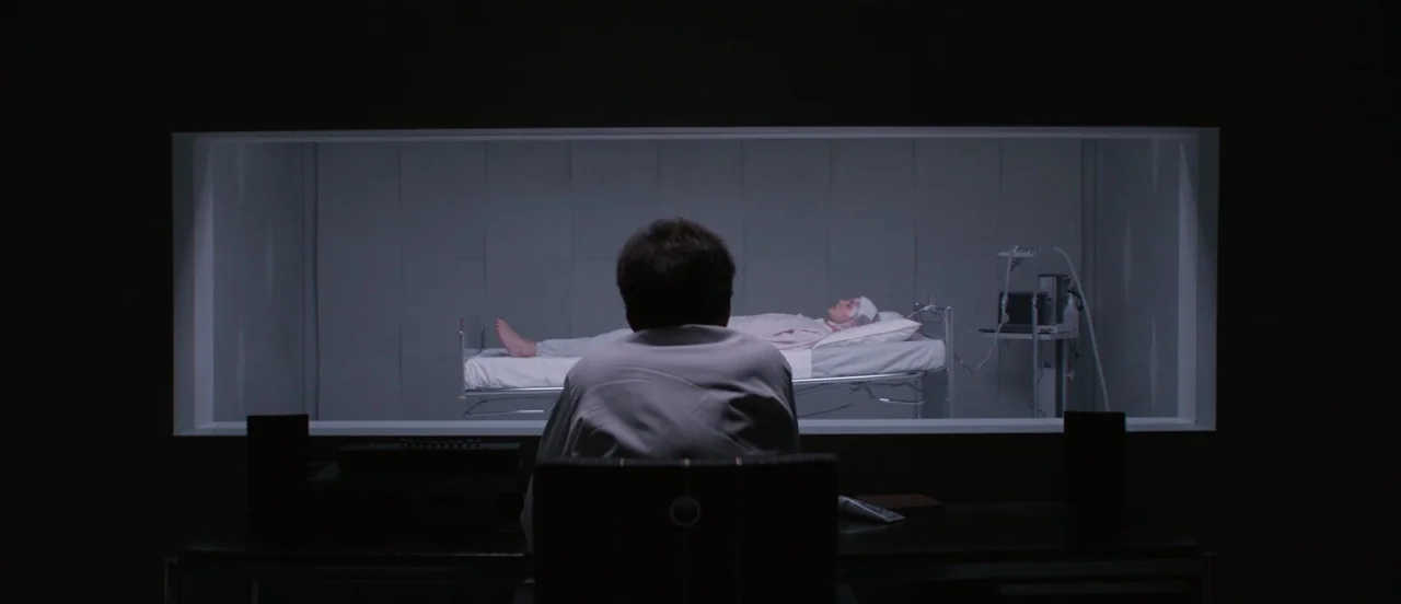 Thelma (2017) movie still: wide — A dark observation room features a large rectangular window looking into a sterile medical ward. A person sits at…