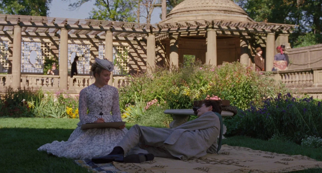 Little Women (2019) movie still: wide — A stone gazebo with a lattice roof stands in a lush, green public park. A woman sketches on a pad while a…