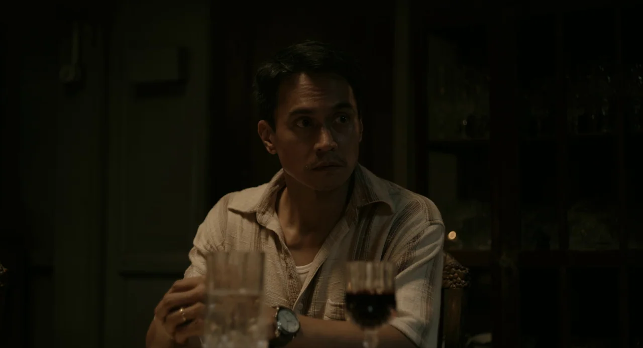 In My Mother’s Skin (2023) movie still: medium — A dimly lit room with dark wooden furniture visible in the soft background. A man sits at a table…