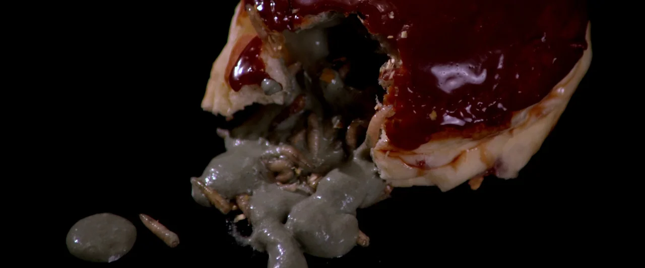 Kaboom (2010) movie still: closeup — A split pastry topped with thick red glaze rests against a solid black background. Maggots and a viscous…
