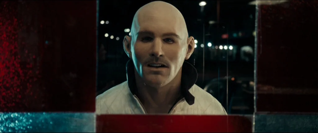 Drive (2011) movie still: closeup — A dark night street is viewed through a checkered glass window pane at night. A bald man wearing a white jacket…