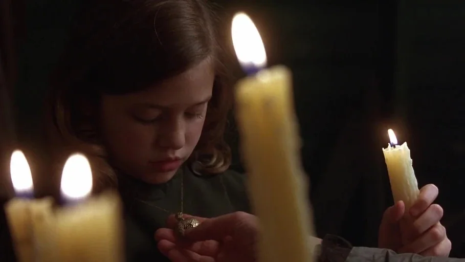A Little Princess (1995) movie still: closeup — A dark, dimly lit room contains a young child with soft brown hair. A hand presents a gold locket to…