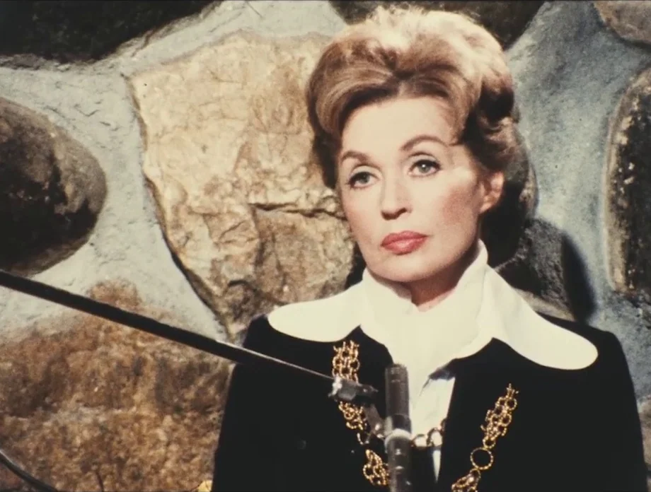 The Other Side of the Wind (2018) movie still: closeup — A woman stands in front of a textured stone wall interior setting. She wears an elegant…