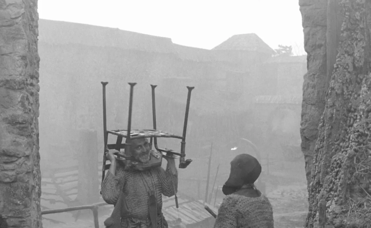 Hard To Be A God (2013) movie still: medium — A misty, desolate village square features stone walls and primitive wooden structures. A man with a…