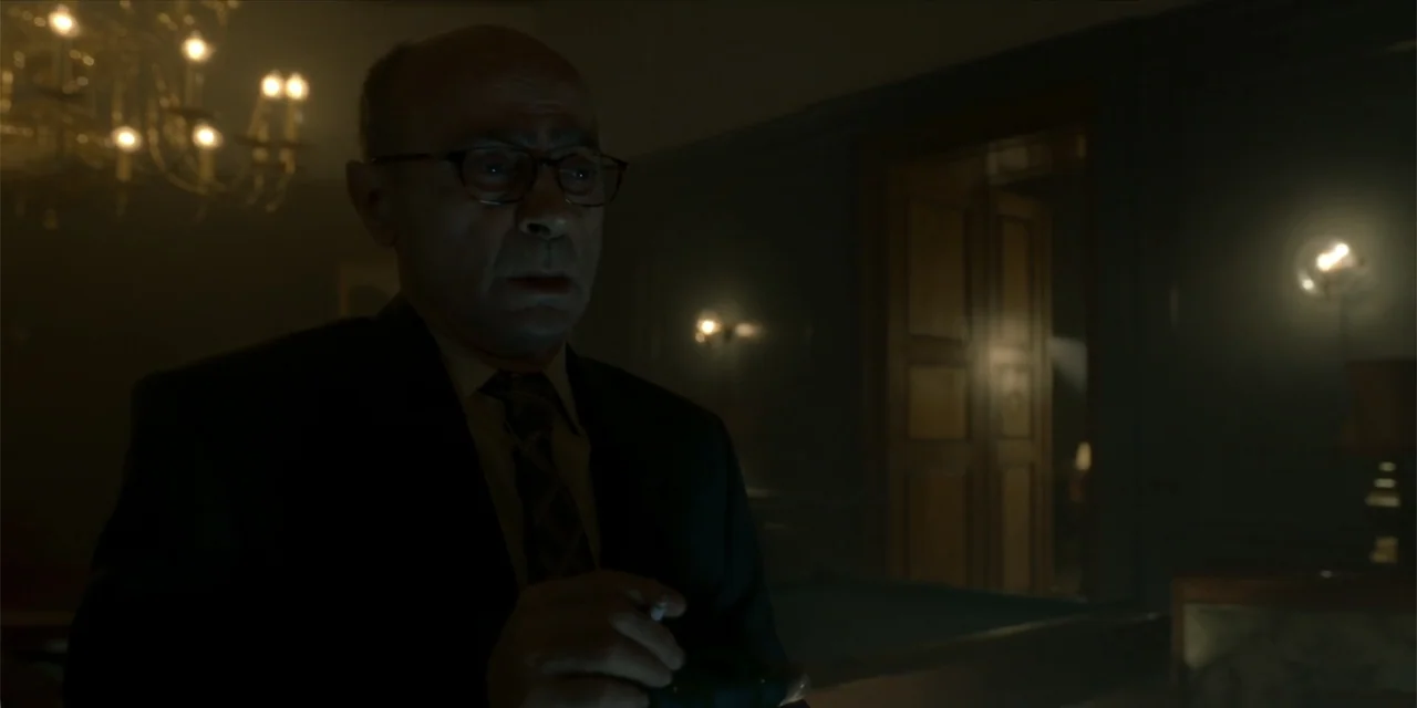 Oslo (2021) movie still: medium — A dark study features a glowing chandelier and a partial view of a doorway. A man in a suit stands in the…