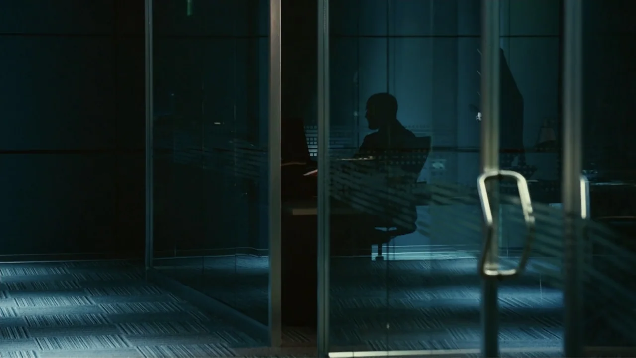 Furious 7 (2015) movie still: medium — A modern glass-walled office space is dimly lit in shades of dark blue. A solitary man sits at a desk while…