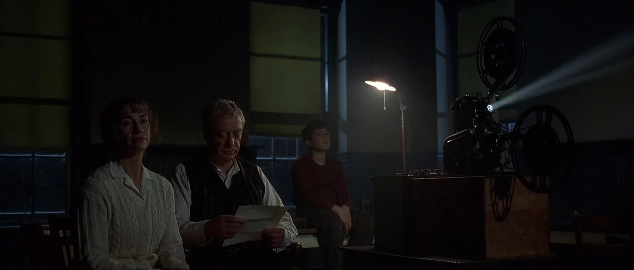 The Cider House Rules movie still: wide — A dark community hall with large windows features a 16mm projector on a table. Three individuals sit in…