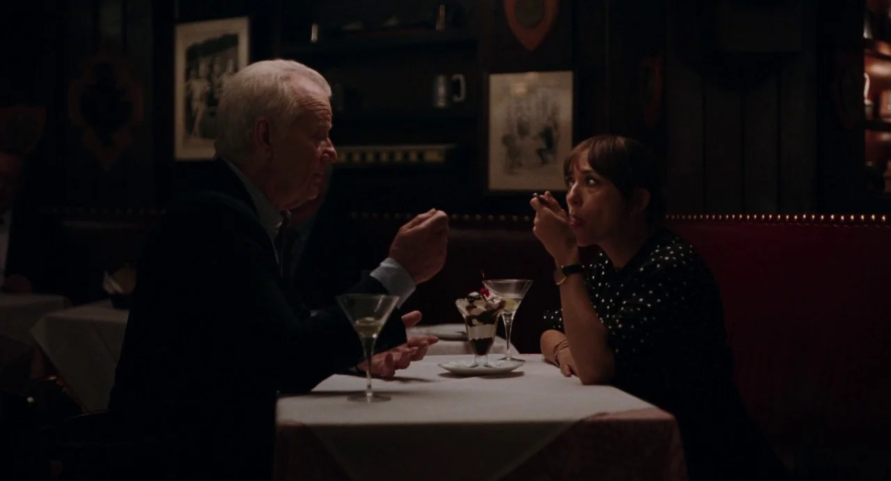 On The Rocks (2020) movie still: medium — An dimly lit booth inside a dark wood-paneled establishment features framed art on walls. A man talks to a…