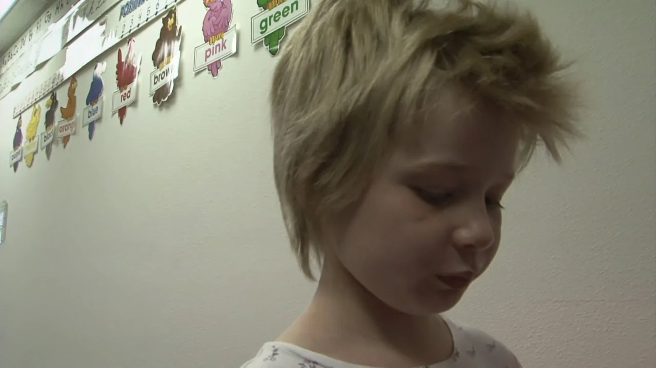 Homeless: The Motel Kids of Orange County (2010) movie still: closeup — A child stands against a white classroom wall decorated with colored word…