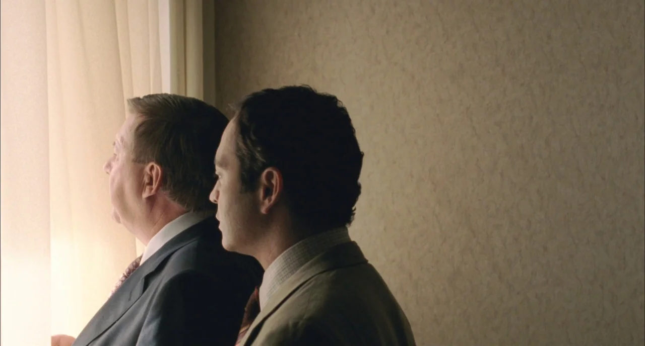 Incendies movie still: medium — A plain interior room with textured beige wallpaper and a white sheer curtain. Two men in business attire stand…