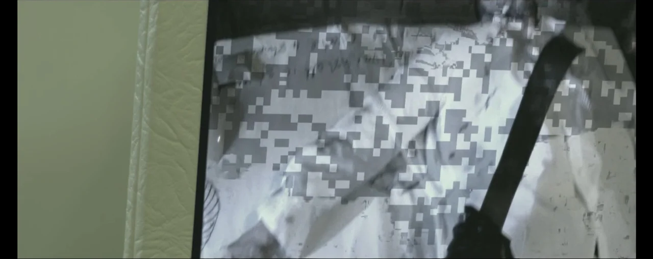 Turbulence (2025) movie still: closeup — A digital screen displays a distorted and pixelated video feed of people. The monitor is encased in a white…