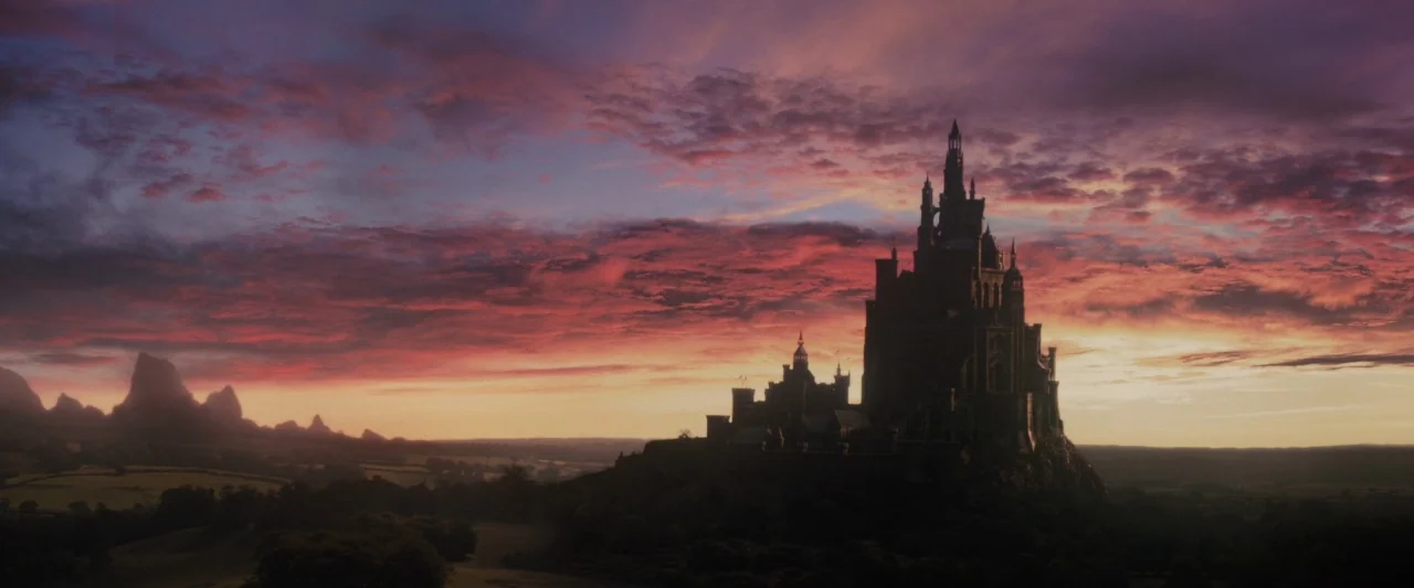 Maleficent (2014) movie still: establishing — A gothic stone fortress occupies a rocky hill in a flat, expansive rural landscape. Vivid sunset…
