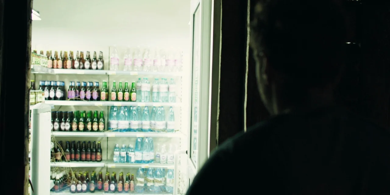 The Ritual (2017) movie still: medium — A person stands in a dimly lit space facing a bright open-front refrigerated display. Rows of various glass…