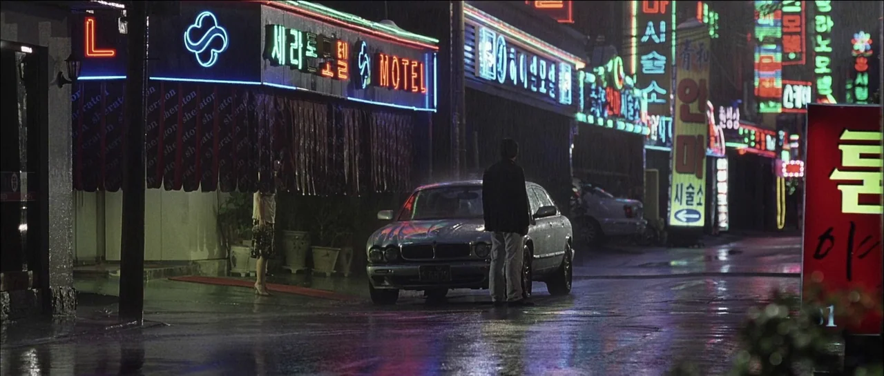 The Chaser (2008) movie still: wide — A wet street at night is lined with glowing multi-colored neon signs in Korean. A man stands behind a parked…