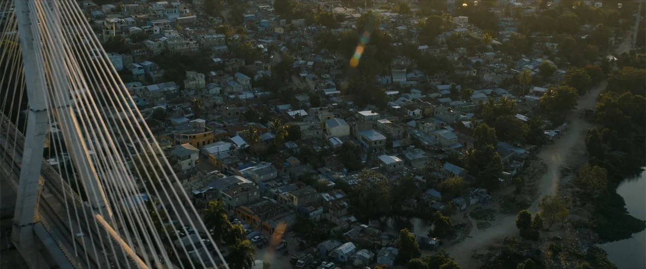 Arthur the King (2024) movie still: establishing — A high-angle view looks down upon a dense collection of small urban dwellings. A large…