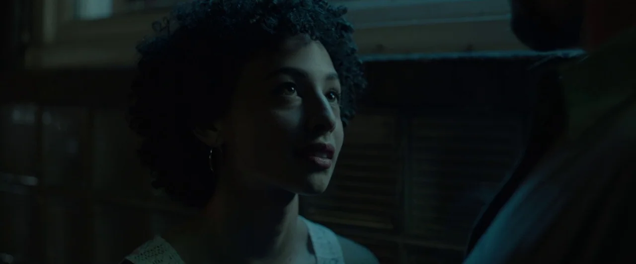 Small Crimes (2017) movie still: closeup — A dark, dimly lit interior space with a person partially in the foreground. A young woman with curly hair…