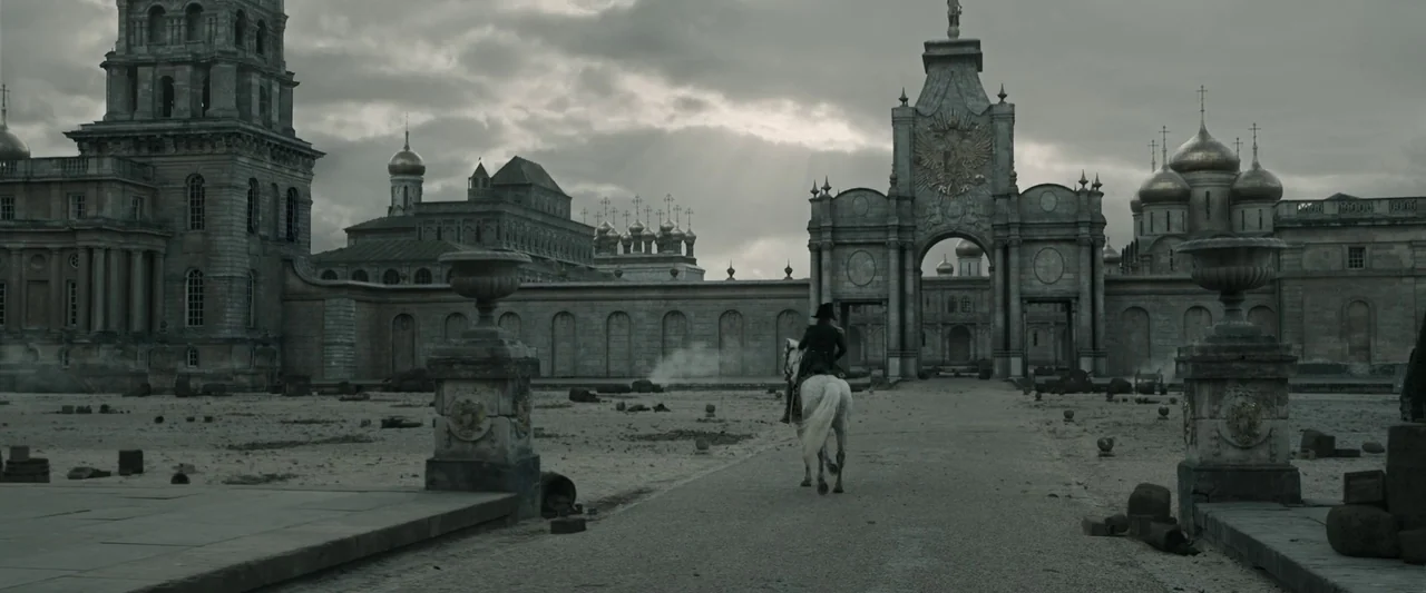 Napoleon (2023) movie still: wide — A vast, desolate stone palace courtyard features a monumental gatehouse under a cloudy sky. A solitary man on a…