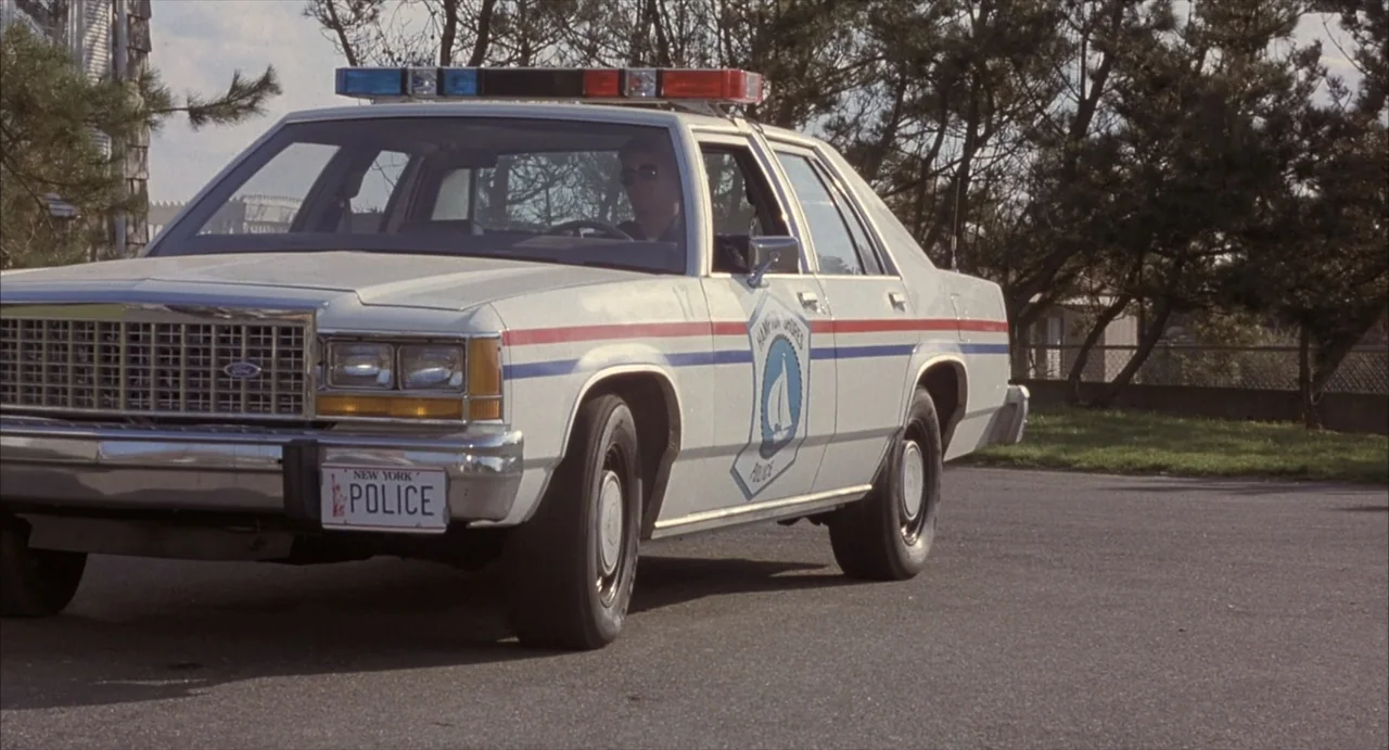 Masquerade (2023) movie still: medium — A white Ford police sedan with red and blue emergency lights parked on asphalt. A man in uniform drives the…