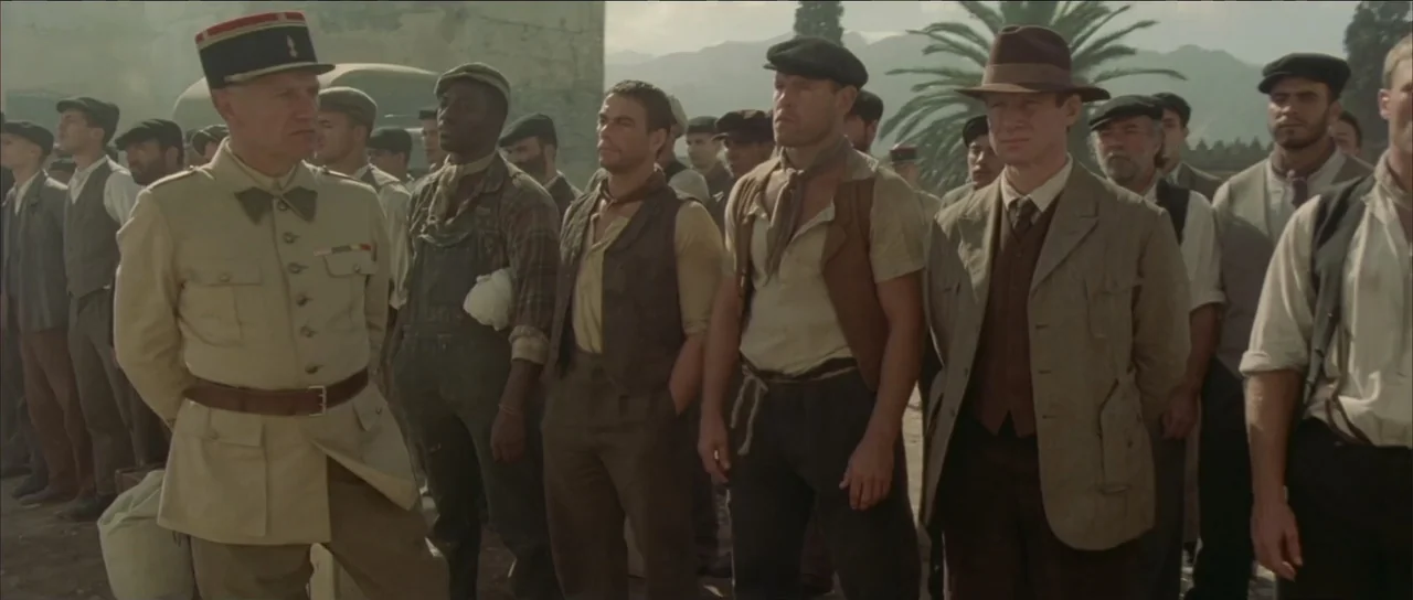 Legionario (2025) movie still: medium — A group of diverse men in 1930s-style clothing stand outdoors in a dusty training area. An officer in…