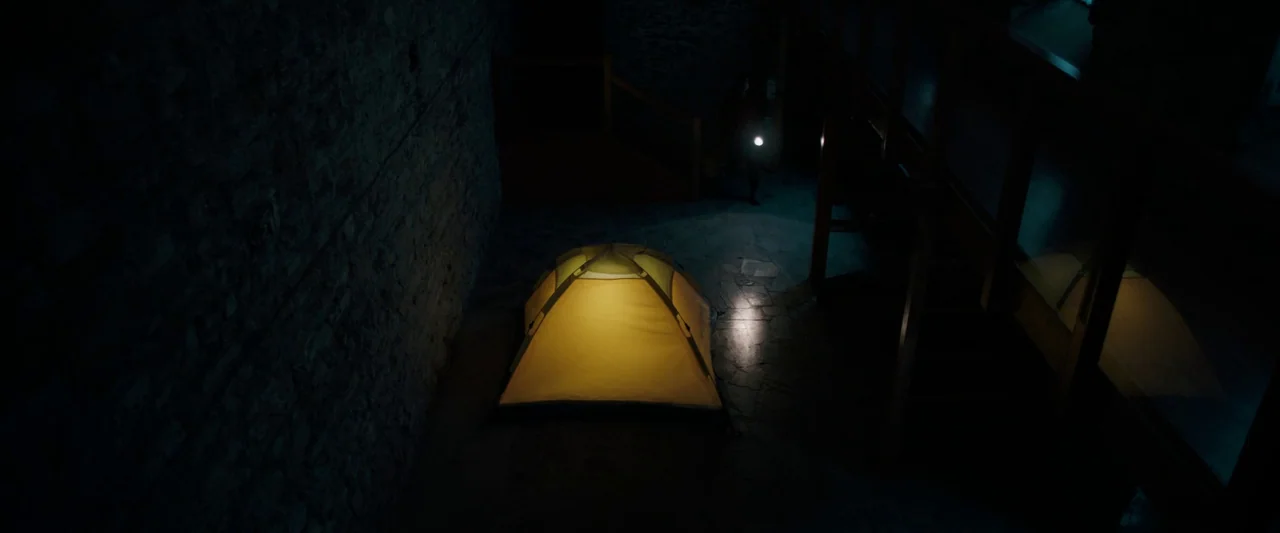 Oddity (2024) movie still: wide — A stone basement room with dark walls contains a brightly lit yellow tent. An empty concrete floor extends from…
