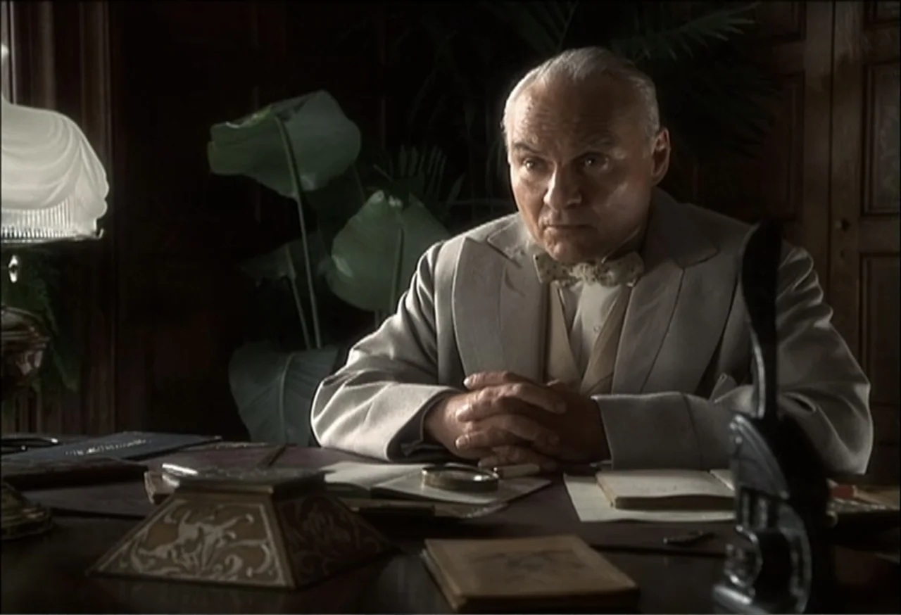 Vendetta (2022) movie still: medium — A dimly lit mahogany-paneled office contains a cluttered desk with a lamp and paperwork. An elderly man in a…