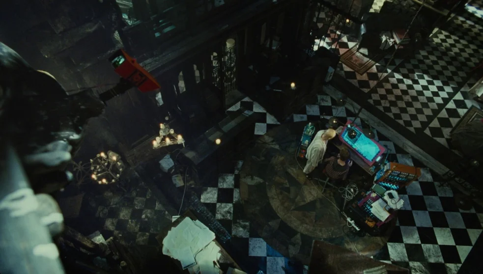 The Zero Theorem (2013) movie still: wide — A gothic-inspired study features a black and white checkered tile floor and dark wooden furniture. Two…