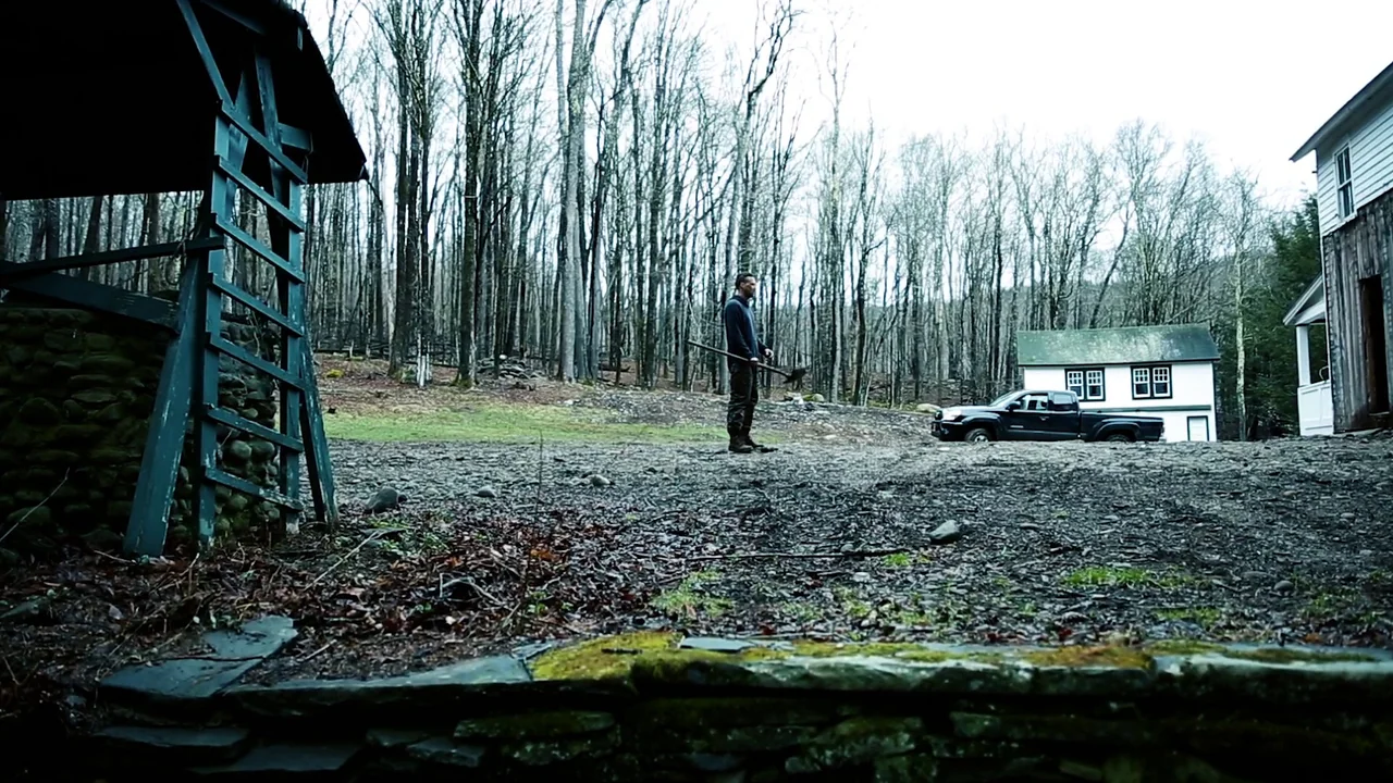 The Deeper You Dig (2019) movie still: wide — A rural lot with a dirt ground leads toward a dense forest of leafless trees. A man holding a…