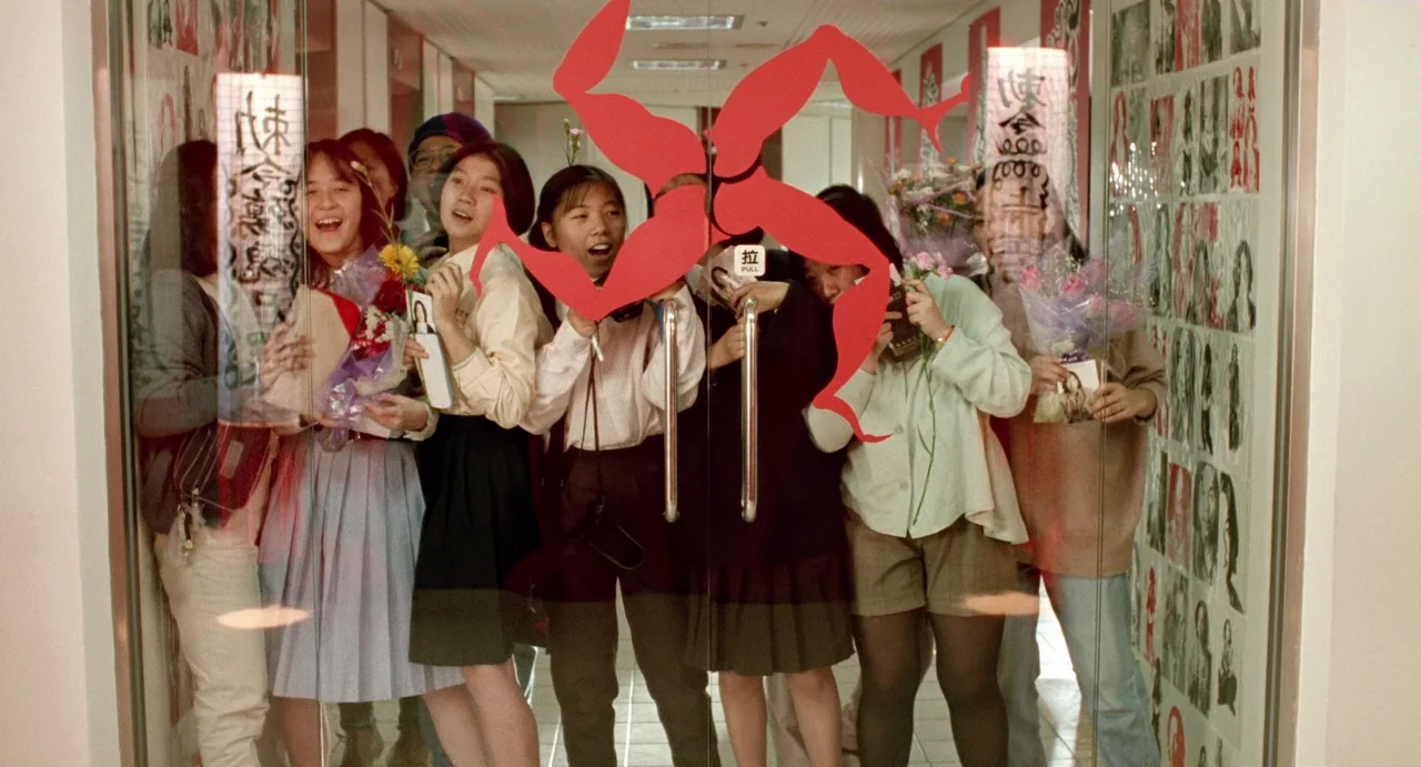 A Confucian Confusion (1994) movie still: medium — A group of young people stand behind a glass door plastered with a red graphic. They hold flowers…