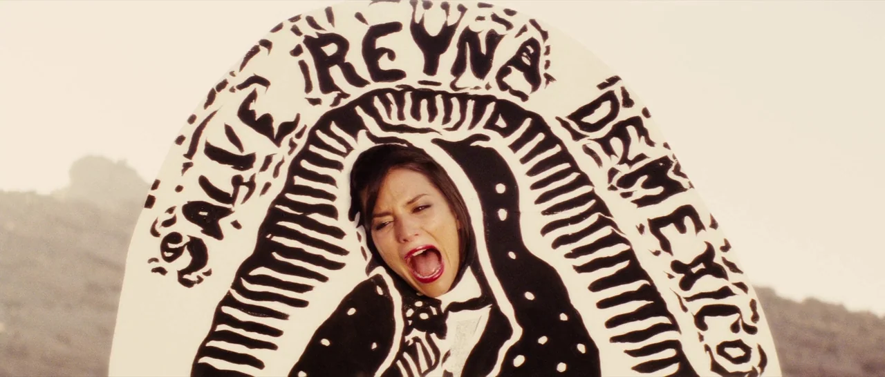 Casa De Mi Padre (2012) movie still: closeup — A woman screams through a cutout in a large, white, religious-themed painted wooden sign. The sign…