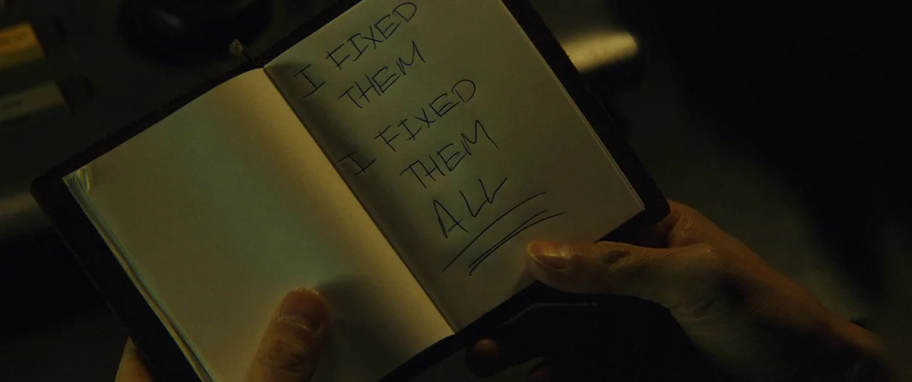 Broadcast Signal Intrusion (2021) movie still: closeup — A small notebook is held open displaying the handwritten message I FIXED THEM ALL. The…