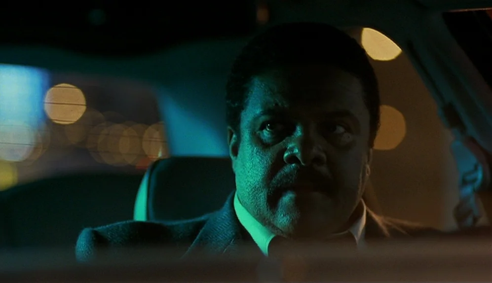 The Limey (1999) movie still: closeup — A man sits in the driver seat of a vehicle at night. Green and orange city lights reflect through the car…