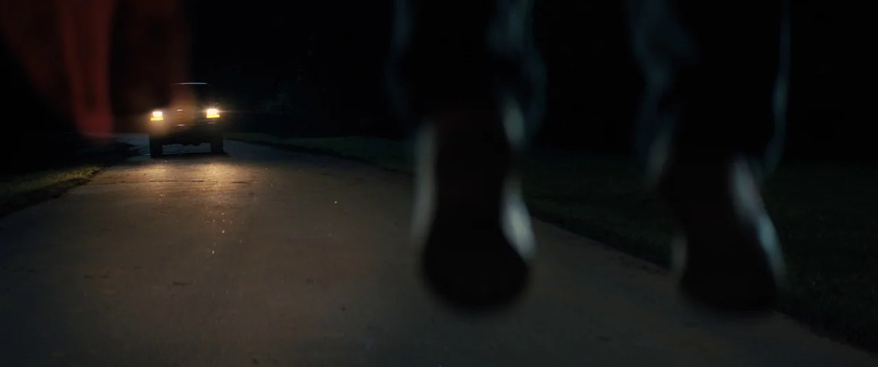 Brightburn (2019) movie still: wide — A dark, empty rural road is illuminated by the oncoming headlights of a vehicle. A person's out-of-focus feet…