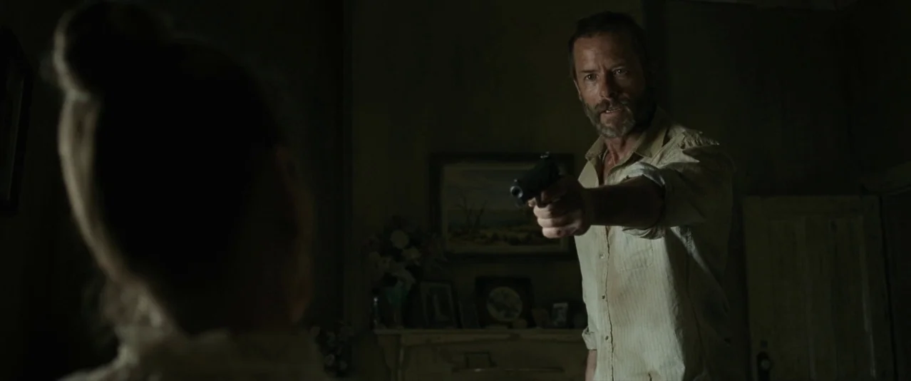 The Rover (2014) movie still: medium — A dimly lit room containing a fireplace mantle decorated with framed photographs and floral arrangements. A…