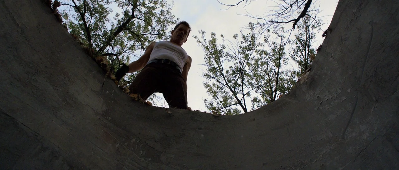 Cop Car (2015) movie still: fullbody — A man stands at the edge of a deep concrete circular shaft outdoors. Looking downward, he is framed from…