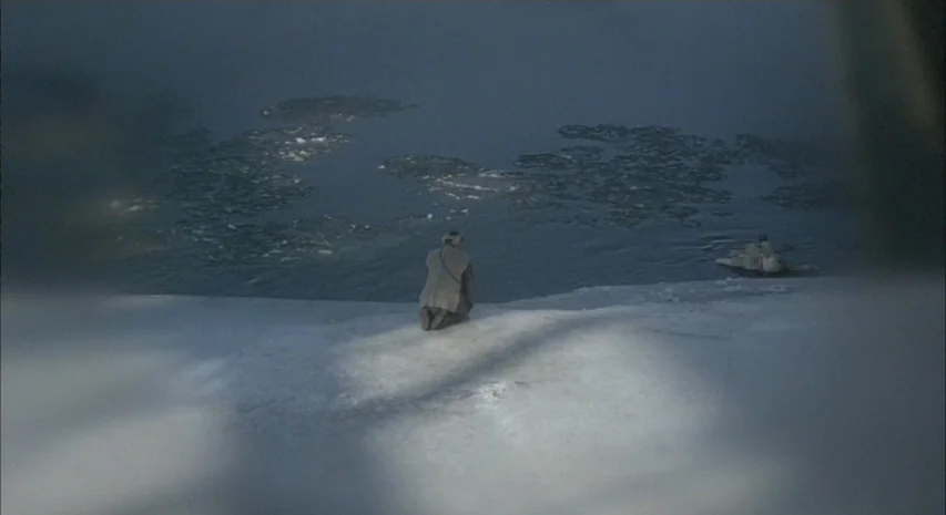 Quintet (1979) movie still: wide — A snowy, icy riverbank under an overcast sky with chunks of ice in water. A solitary person in a wool coat kneels…