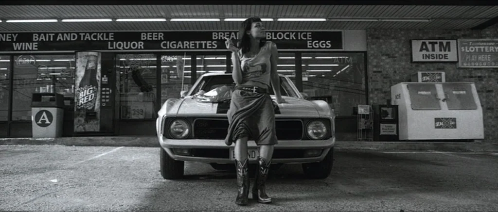 Death Proof movie still: fullbody — A rural store exterior at night with signage advertising bait, tackle, and beer. A young woman leans against the…