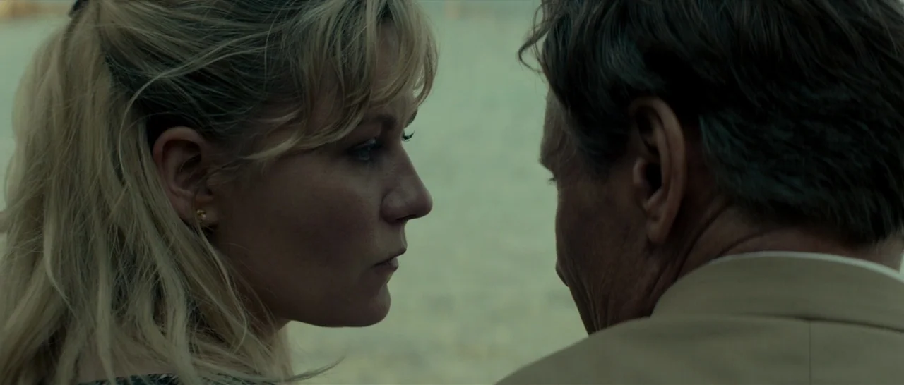 The Two Faces of January (2014) movie still: closeup — A woman and an older man stand facing one another on a sandy shore. The two subjects occupy…