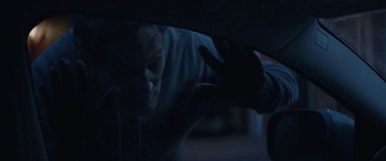 Pet Sematary (2019) movie still: medium — A man leans towards the driver side window of a car at night. His palms and fingers are pressed against…