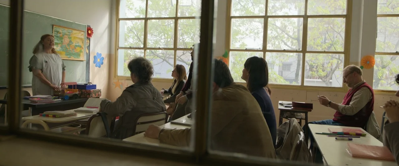 Doublespeak (2021) movie still: wide — An interior classroom features rows of wooden desks and large windows facing exterior trees. A group of adult…