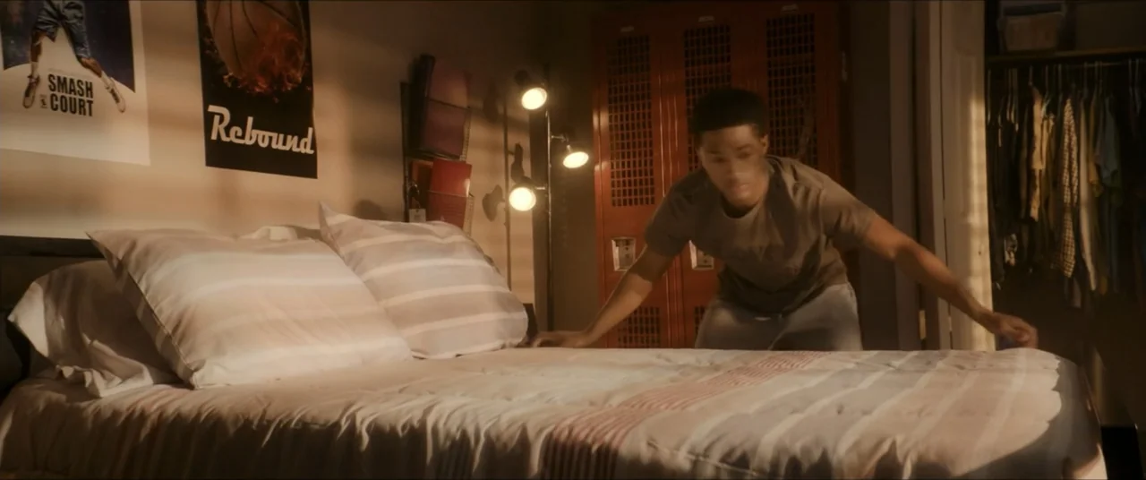 The Forge (2024) movie still: medium — A teenager's bedroom features a bed with striped linens and red lockers. A young man reaches across the…