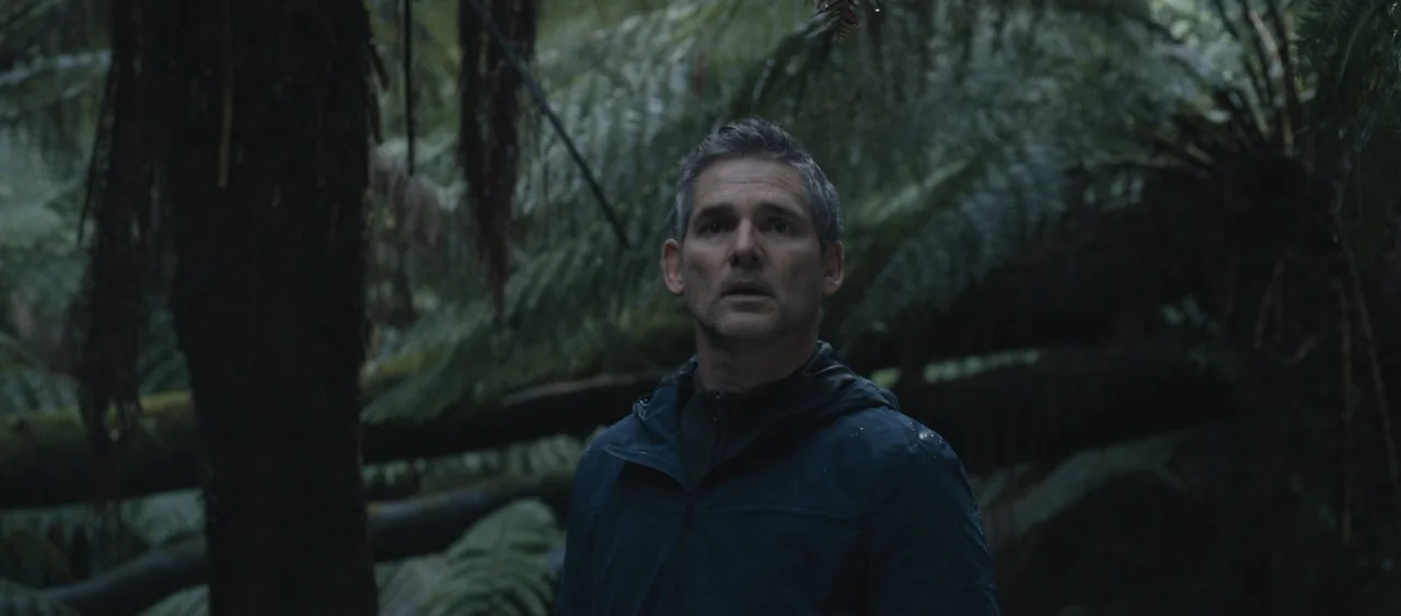 Force of Nature: The Dry 2 (2024) movie still: medium — A dark, wet rainforest environment features dense tree trunks and lush fern foliage. A man…