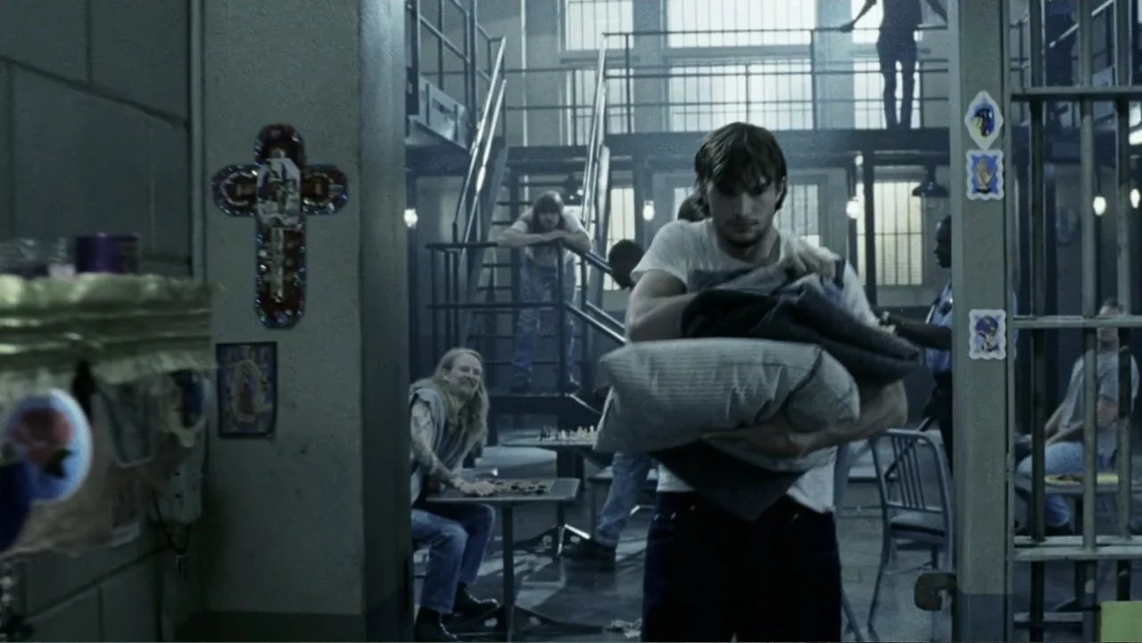 The Butterfly Effect (2004) movie still: medium — A prison common area with steel railings and tables in the background. A young man carries a stack…