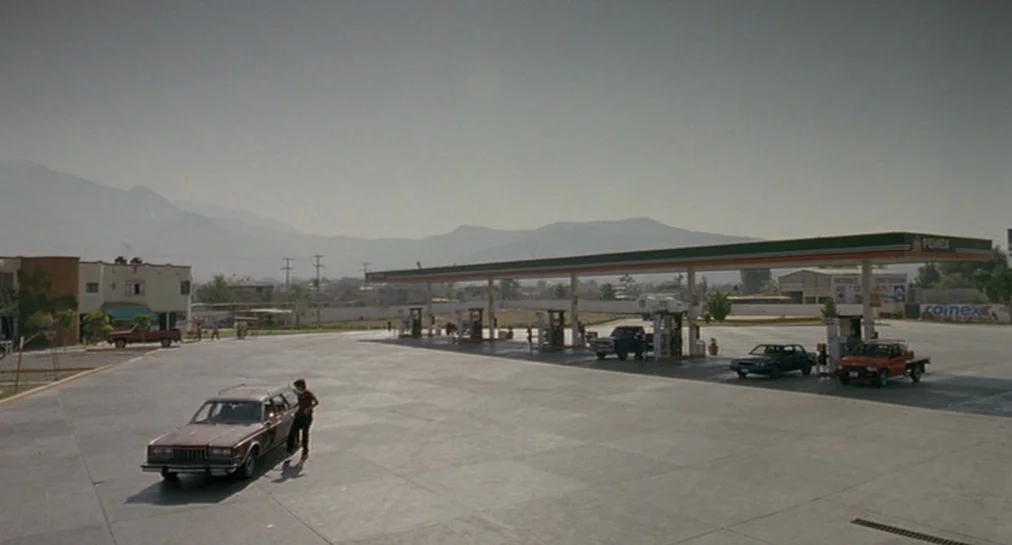 Y Tu Mamá También (2001) movie still: wide — An expansive, paved gas station lot sits against a backdrop of distant mountains. A lone individual…