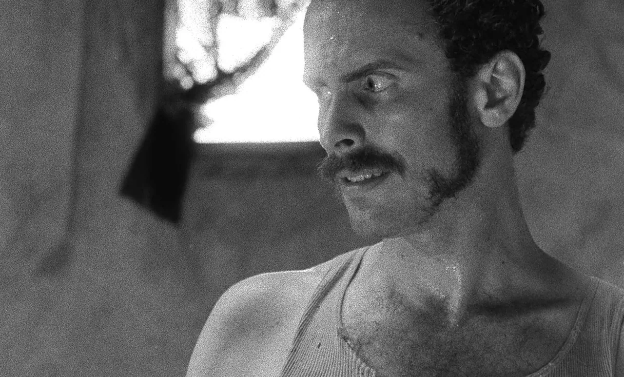 Go Down Death movie still: closeup — A dimly lit room features a small window with a dark curtain. A man with a mustache and curly hair looks…