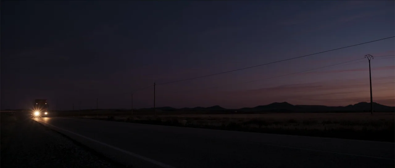 Historias lamentables (2021) movie still: wide — A vast, dark rural landscape with a long road stretching toward distant rolling hills. A single…